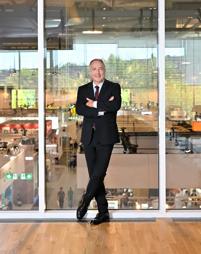 A portrait of Oliver P. Kuhrt, Managing Director of MESSE ESSEN, in front of a glass façade overlooking the exhibition hall.